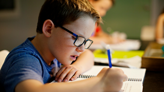A young boy wearing glasses is sitting at a desk writing in a notebook.